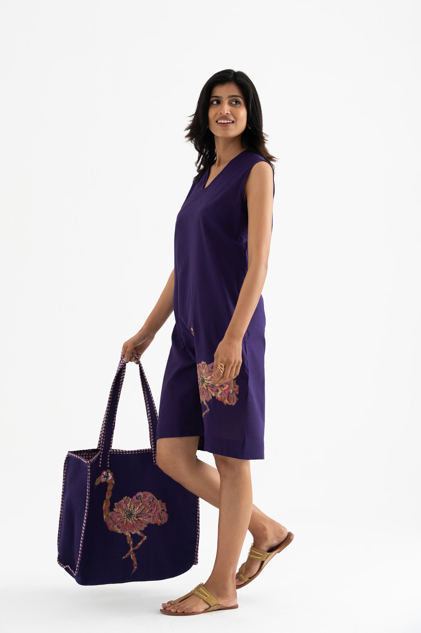 A woman standing, wearing a sleeveless deep purple dress with a V-neckline, paired with a matching large tote bag featuring a block-print flamingo pattern.