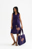 A woman standing, wearing a sleeveless deep purple dress with a V-neckline, paired with a matching large tote bag featuring a block-print flamingo pattern.