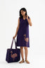 A woman standing, wearing a sleeveless deep purple dress with a V-neckline, paired with a matching large tote bag featuring a block-print flamingo pattern.